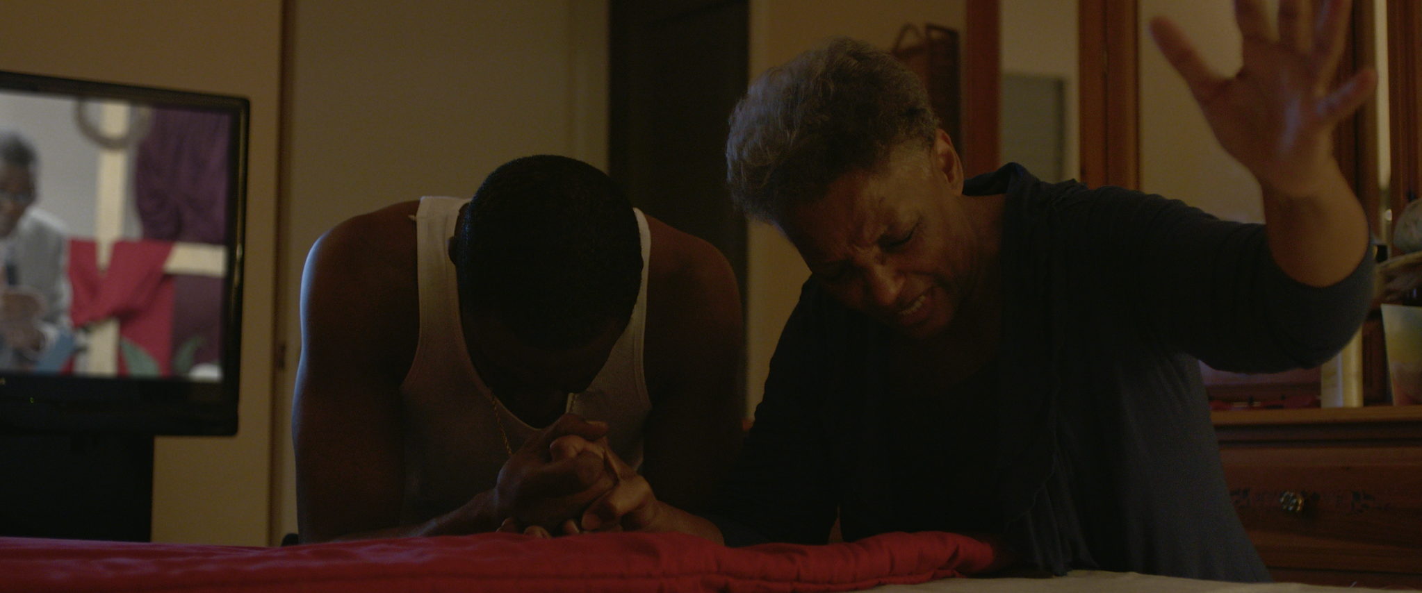 Two people bend over a bed with hands clasped in prayer, a television glowing behind them.