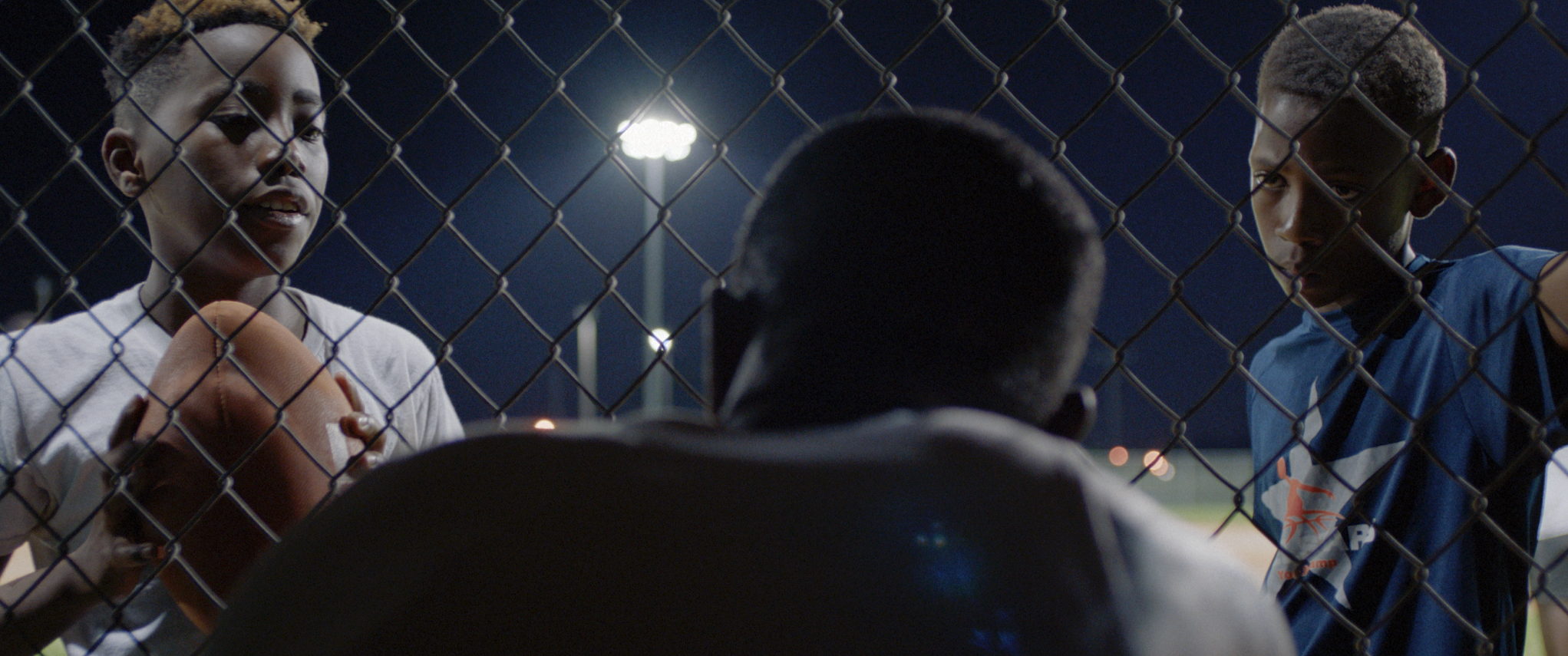 Seen through chain-link at night, a group of men stand talking under a street light.