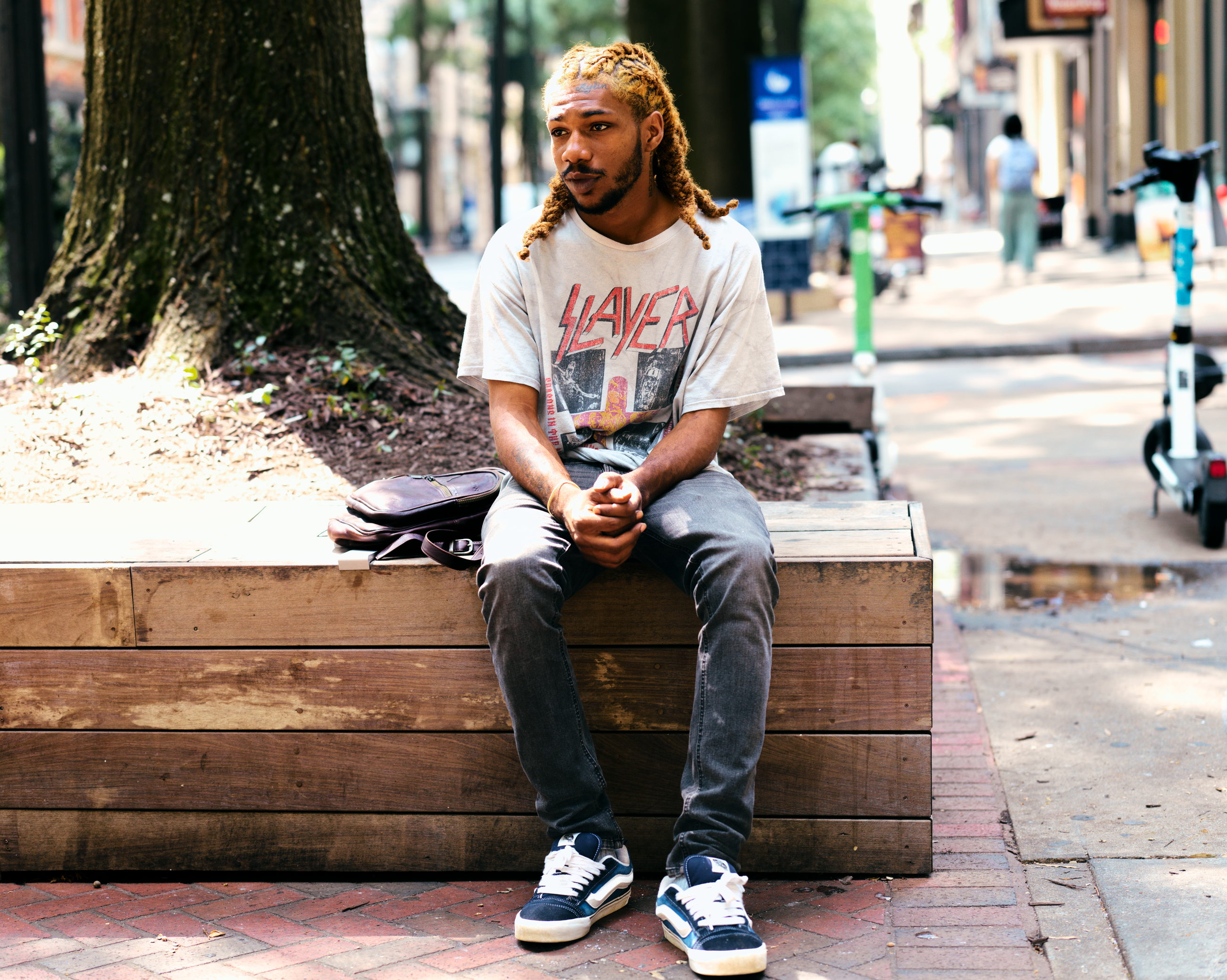 A young man sits on a wooden bench on a city sidewalk, hands clasped between his knees, looking away past the camera; parked scooters and passers-by recede behind him.