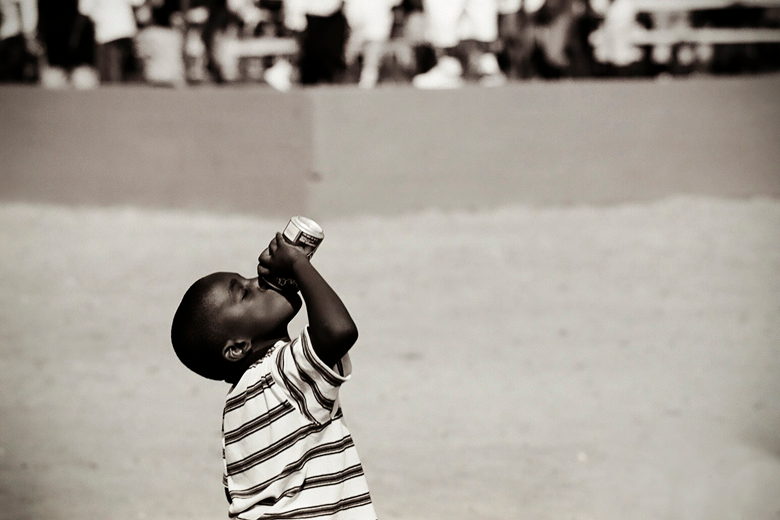 A small boy tips a bottle back to drink, a blurred crowd behind him.