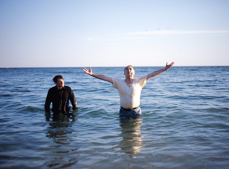 Two men stand waist-deep in the ocean; one opens his arms wide to the sky.